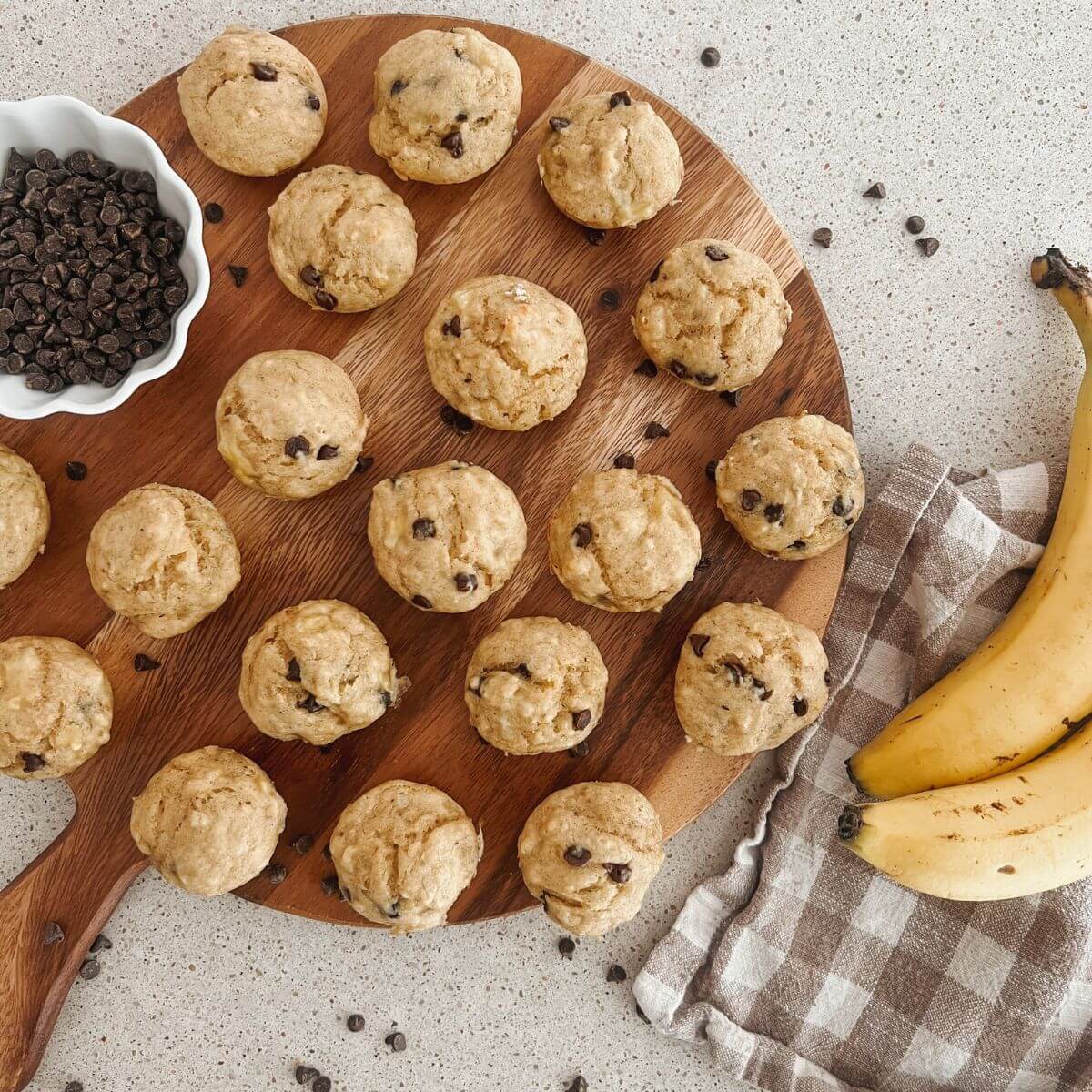 Banana Greek Yogurt Mini Muffins on a cutting board with mini chocolate chips in a white bowl and bananas and a checkered bowl next to it.