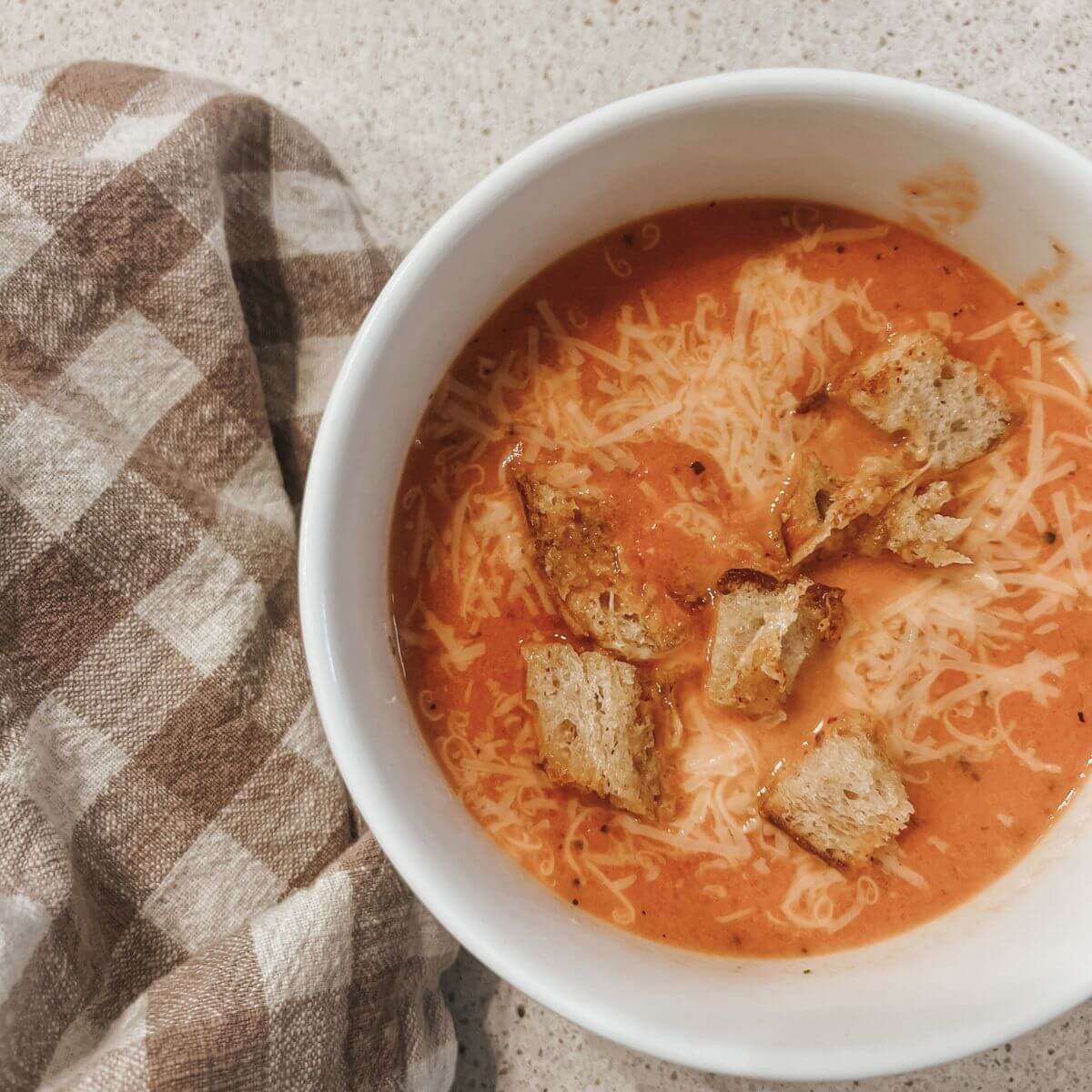 Creamy Tomato Soup Recipe in a white bowl topped with parmesan croutons and parmesan cheese with a brown and white checkered towel.