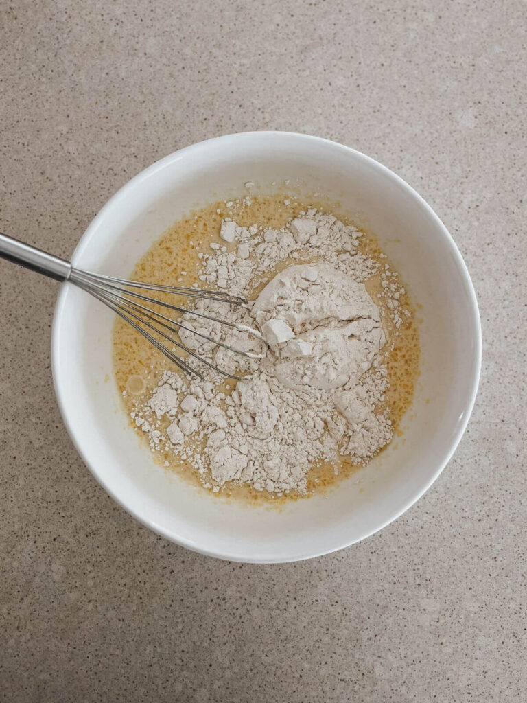 adding the dry ingredients into the wet ingredients for the pancakes in a white bowl with a stainless steel whisk