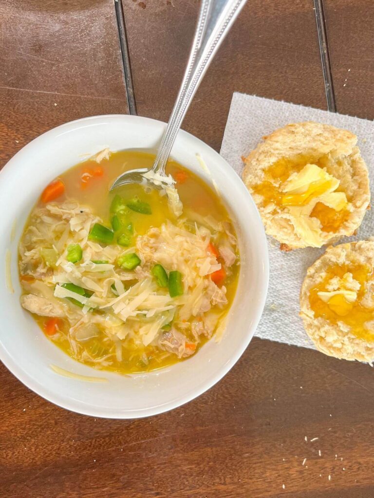 homemade chicken soup in a white bowl with spoon and biscuit next to it. 
