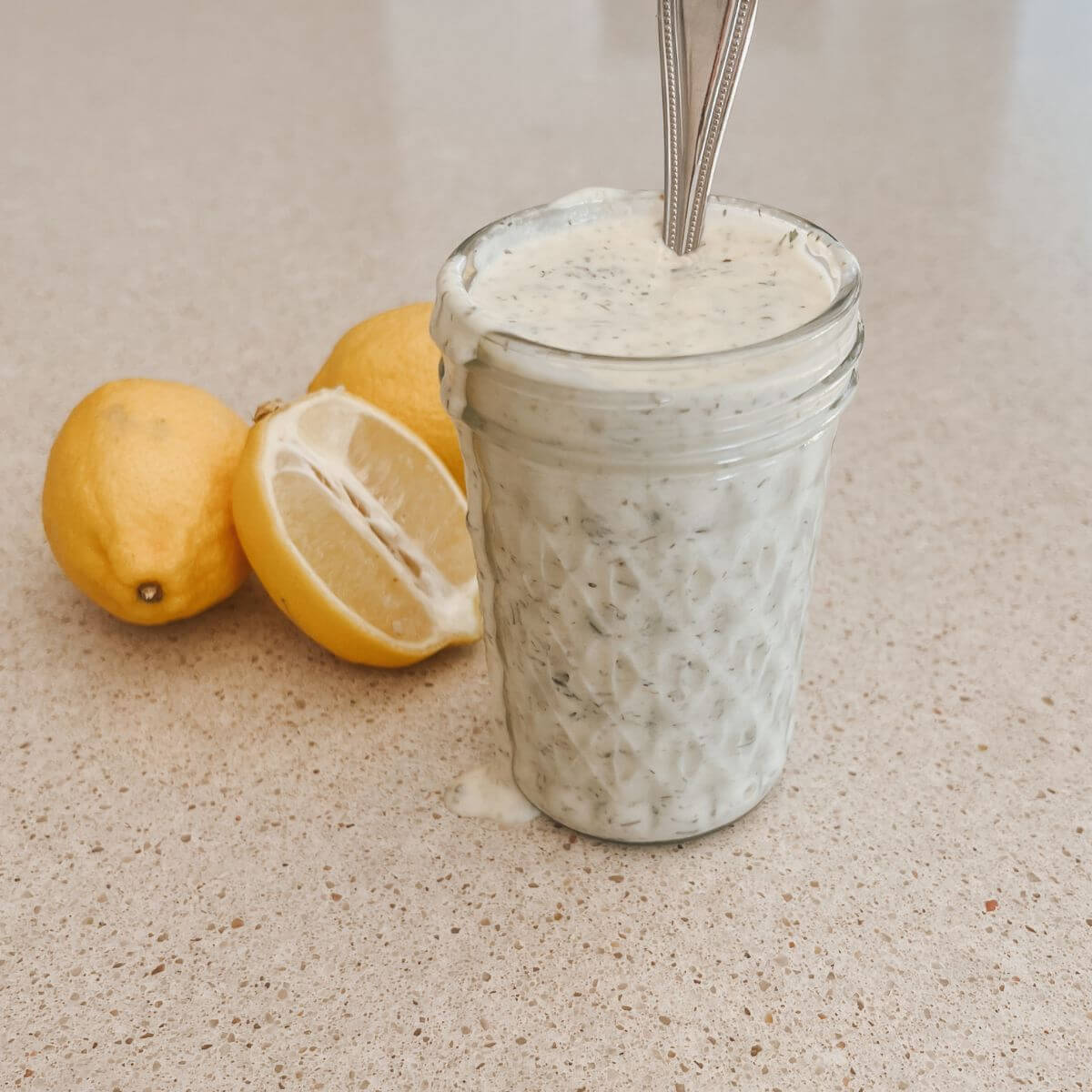 Homemade Ranch dressing in a mason jar with a spoon sticking out on a counter with lemons next to it.