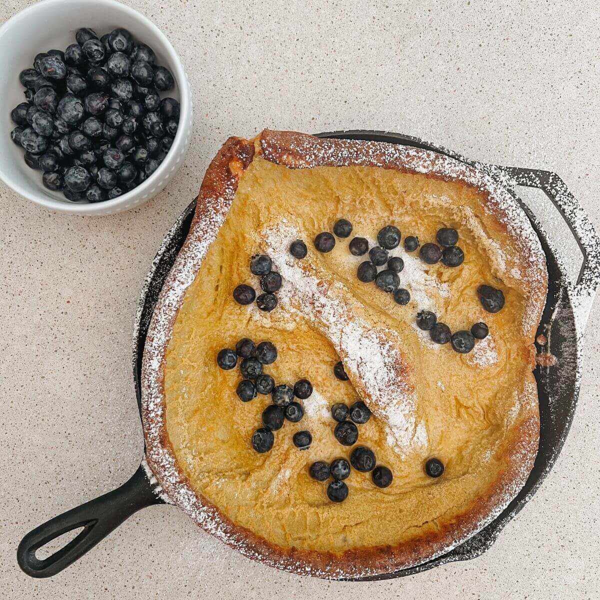 Dutch Baby Pancake in a Cast-Iron Pan topped with powdered sugar and blueberries with a side of blueberries in a white bowl.