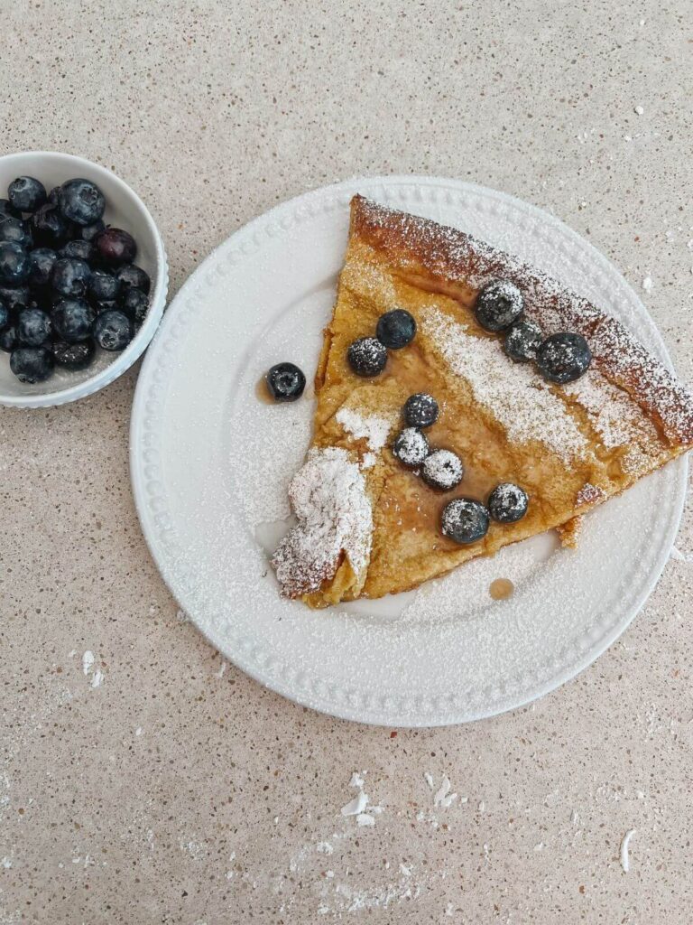 a slice of the pancake on a white plate topped with powdered sugar and blueberries with a side of blueberries in a small white bowl