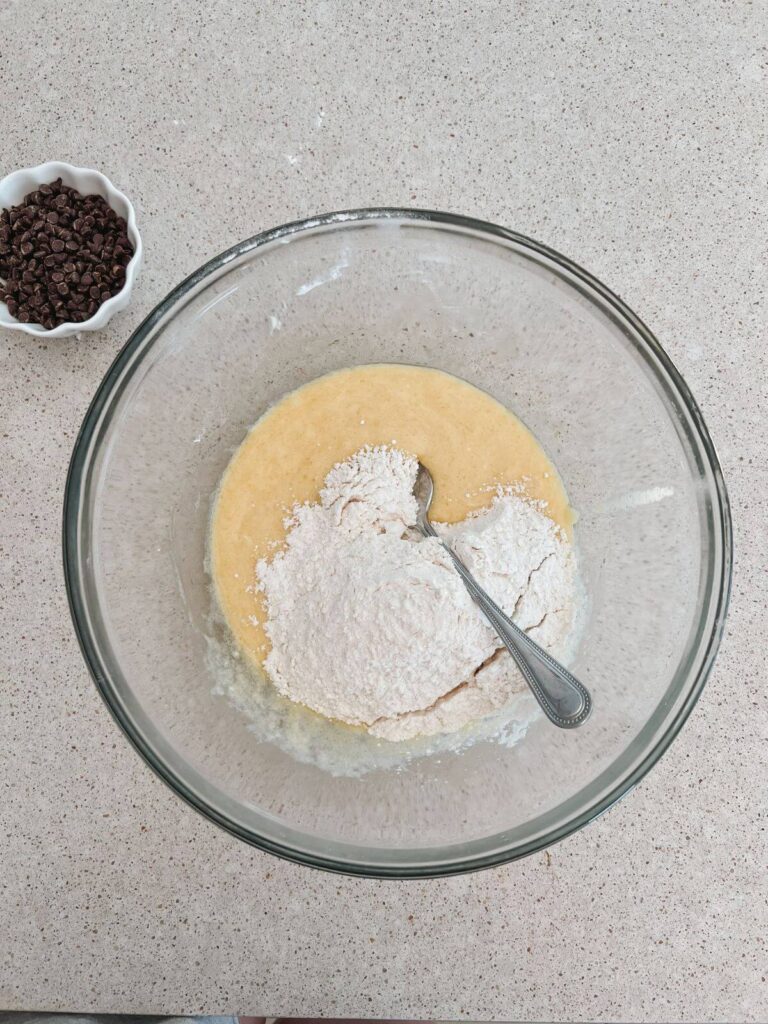 Mixing the wet and dry ingredients together in a glass bowl with a spoon. Chocolate chips are sitting to the side in a small white bowl.