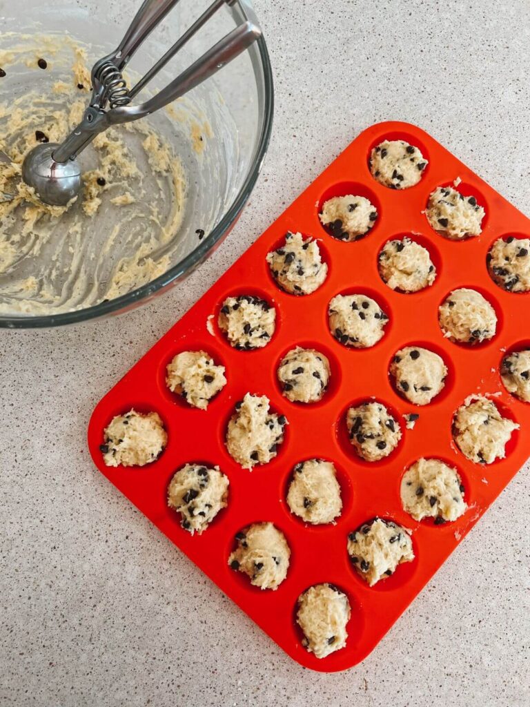 Mini muffin batter in the silicone muffin tin with a glass bowl with the rest of the batter to the side with an ice cream scoop. 