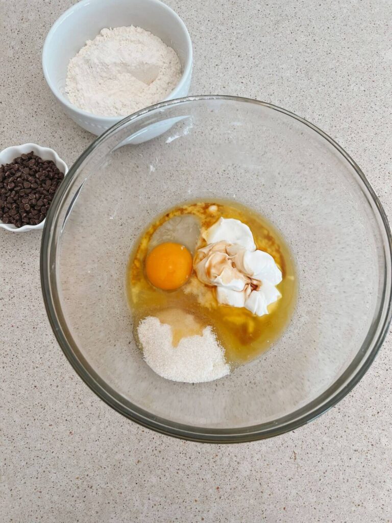 the wet ingredients in a glass bowl with the mixed dry ingredients behind them and the chocolate chips. 