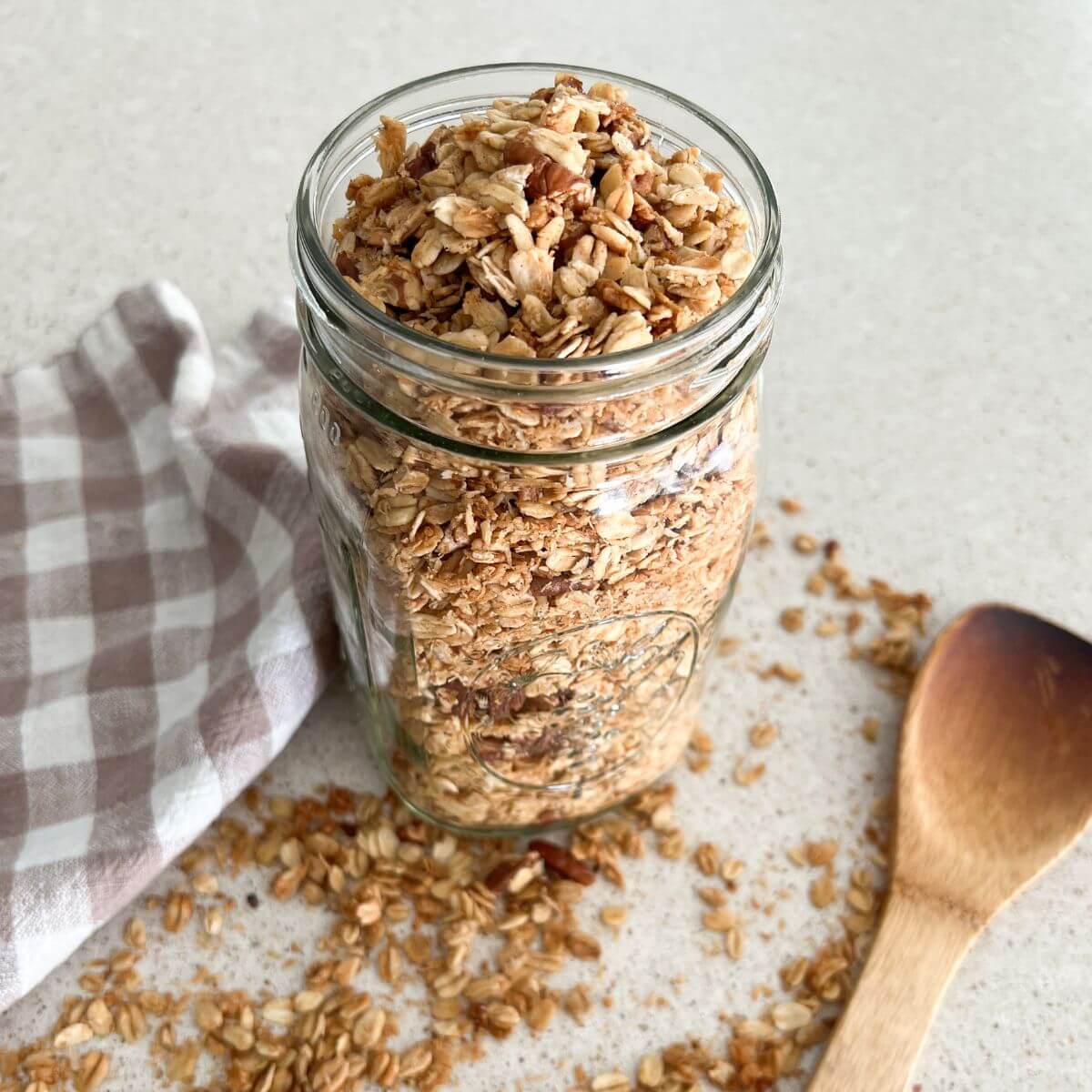 homemade granola in a mason jar with a plaid towel and wooden spoon next to it with granola sprinkled on table.