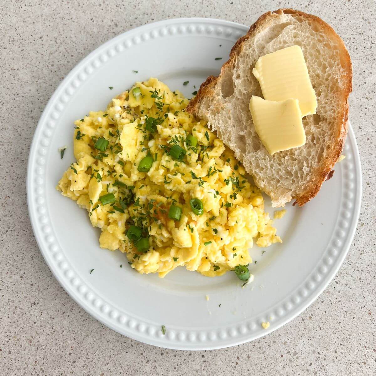 Fluffy scrambled eggs on a white plate with parsley and chives on top with a piece of sourdough toast with butter.