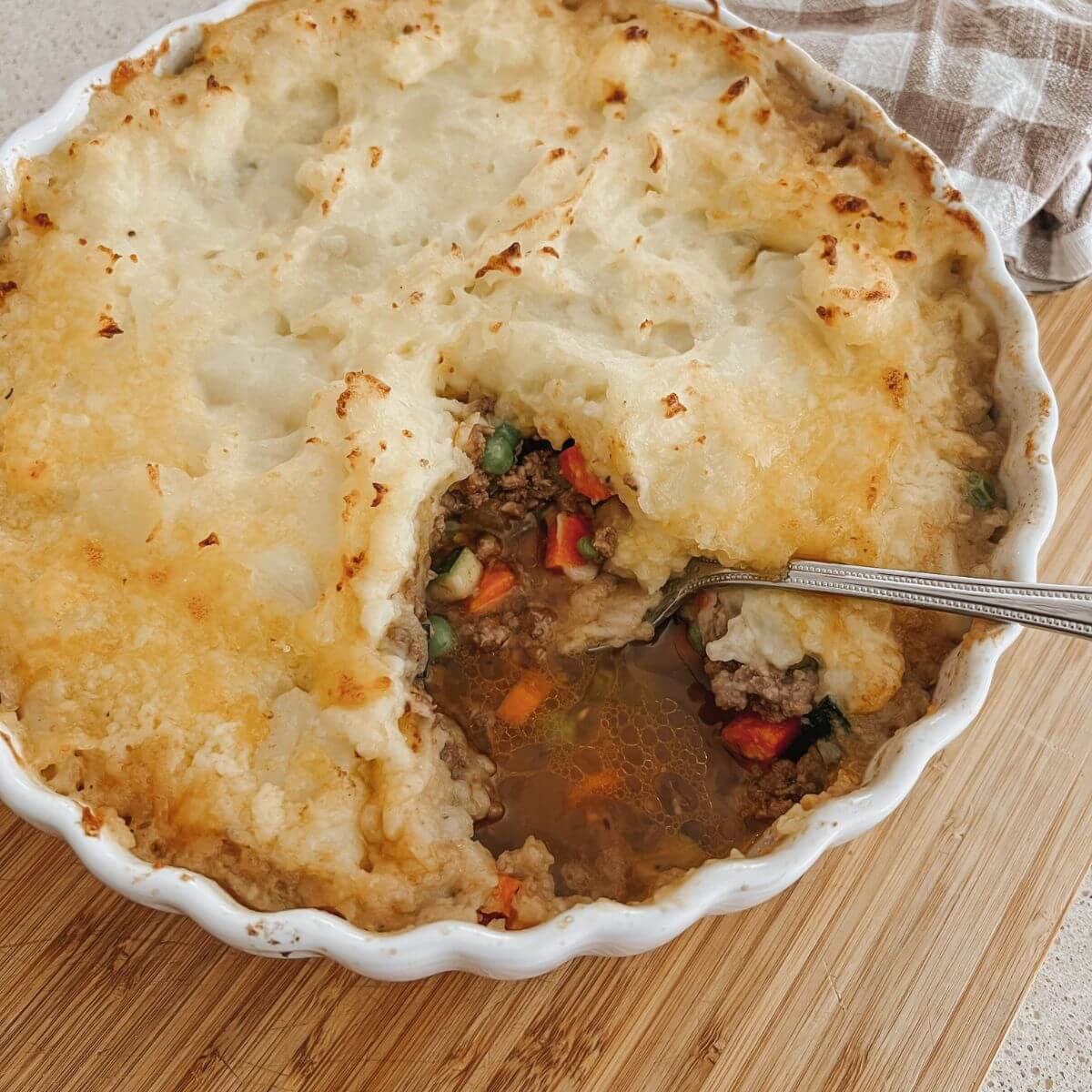 Nourishing Shepherd's Pie in a white round serving dish with a piece cut out with a silver spoon on a wooden cutting board with a plaid towel.