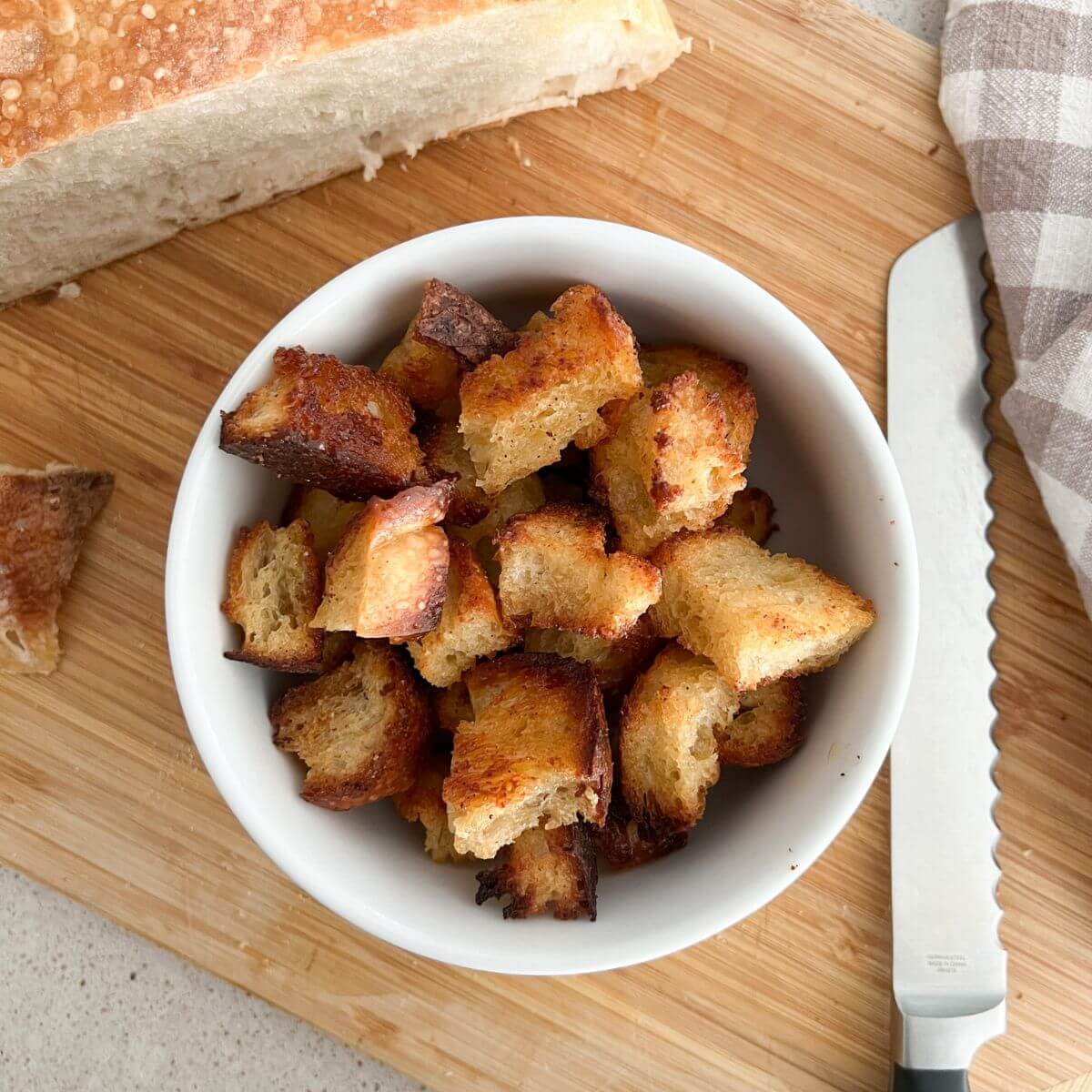 buttery garlic sourdough croutons in a white bowl on a cutting board with a bread knife, plaid bowl, and extra bread next to it