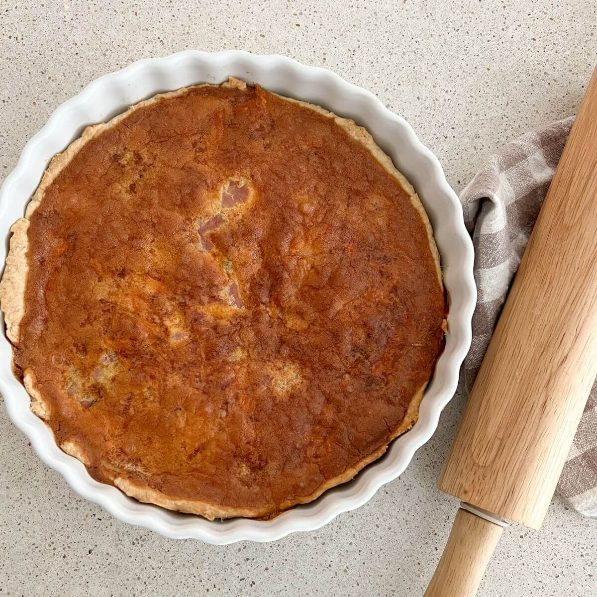 ham and cheese quiche in a white pie pan with a rolling pin and plaid towel next to it