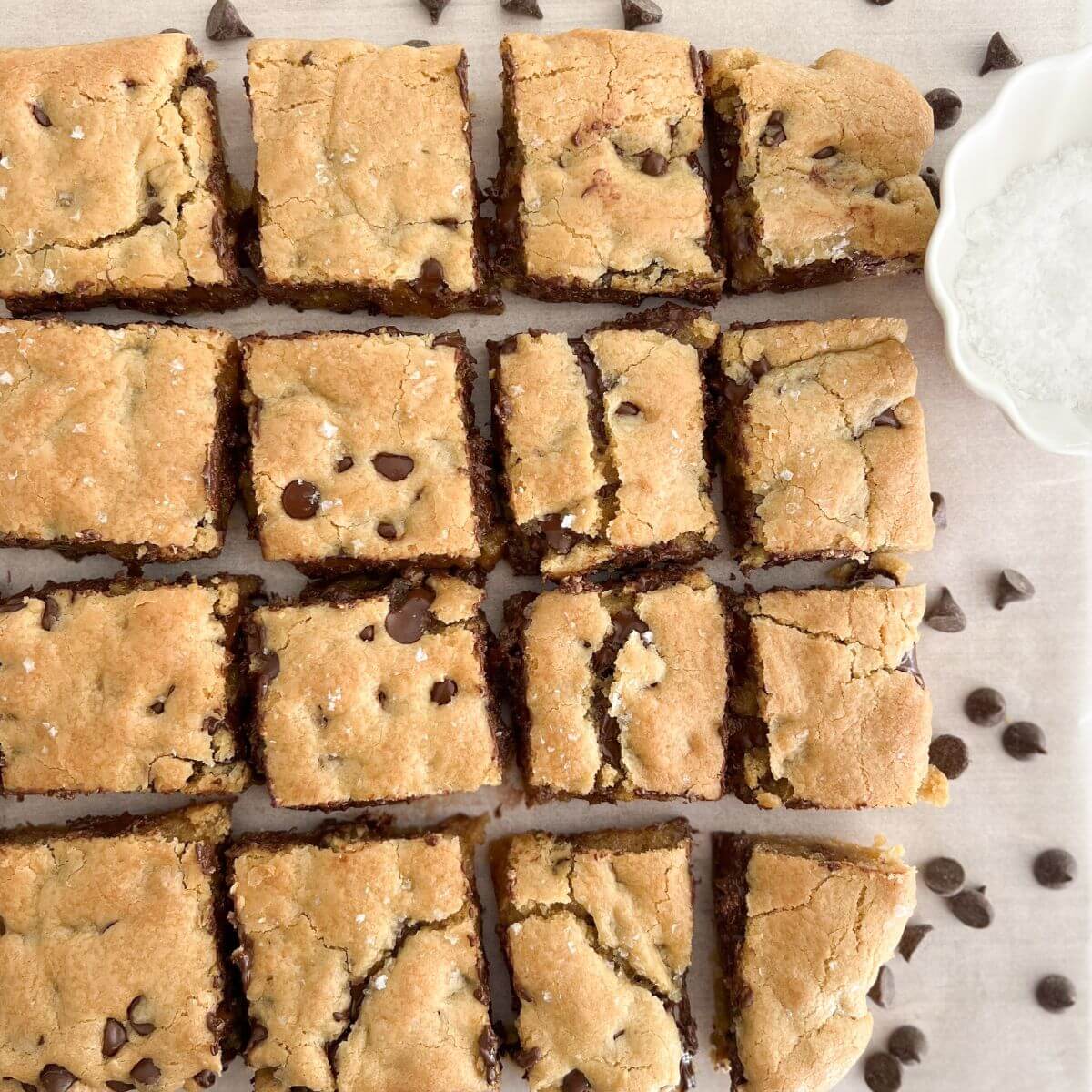 Gooey Chocolate Chip Cookie Bars cut into squares on a piece of white parchment paper with chocolate chips scattered around and a small white bowl with flakey salt.