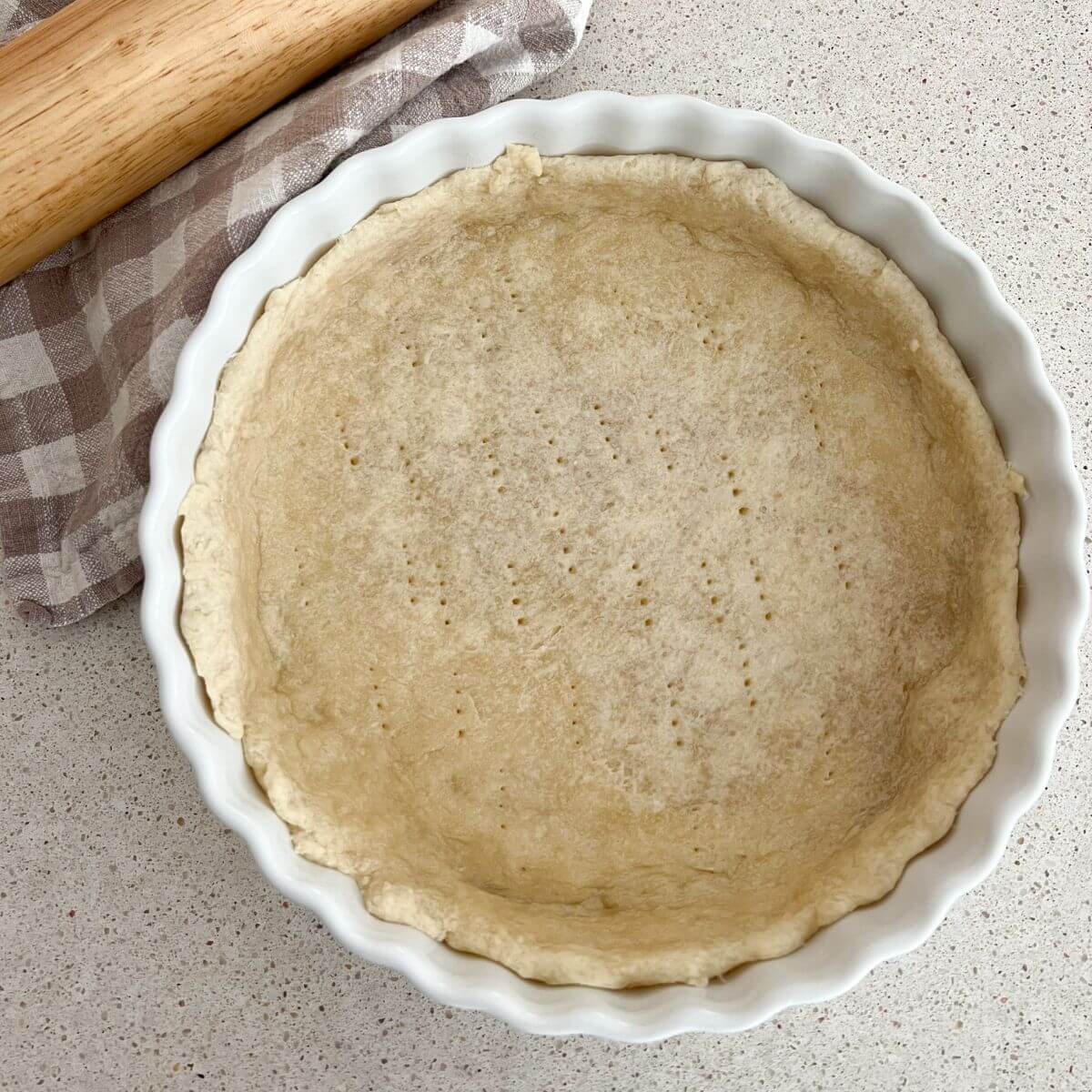 Quick and Easy Pie Crust baked in a white pie pan with a plaid towel and rolling pin next to it