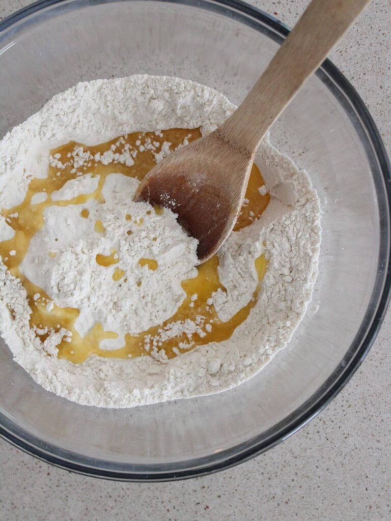 Melted butter being mixed into the flour mixture with a wooden spoon in a glass bowl. 