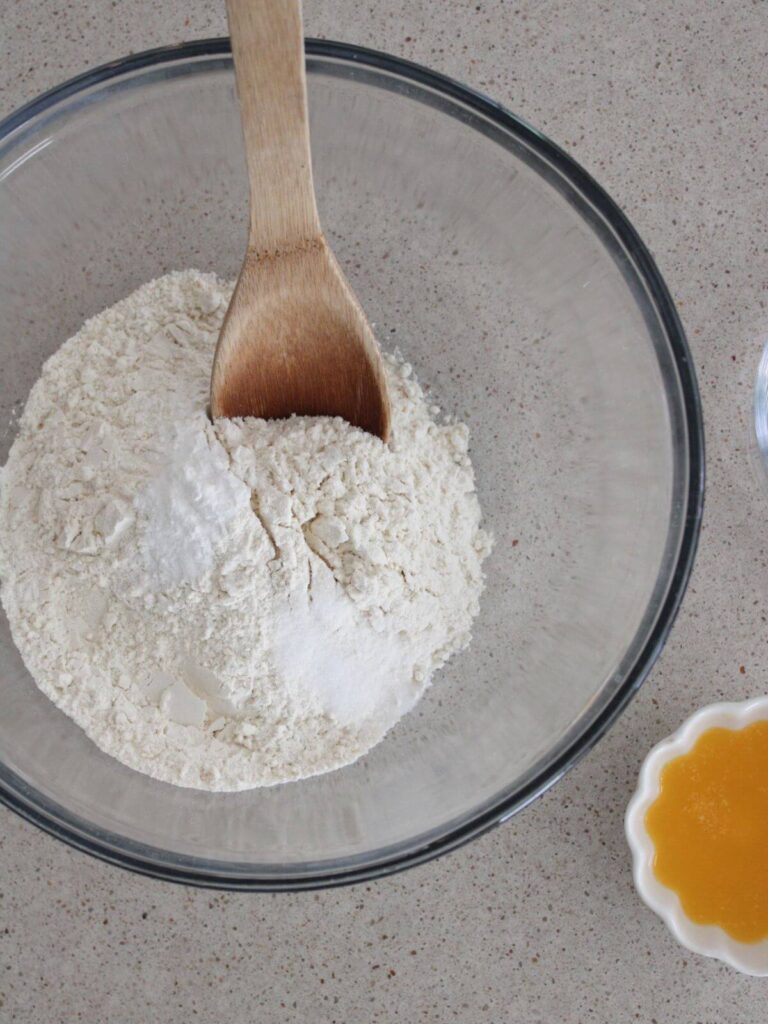 Mixing the flour, baking powder and salt in a large glass bowl with a wooden spoon. Melted butter next to it in a white bowl. 
