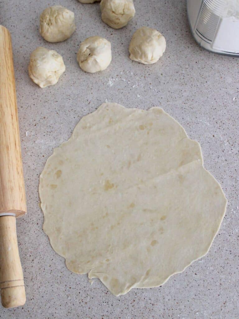 Dough ball rolled out into a circle with rolling pin next to it and other dough balls around.