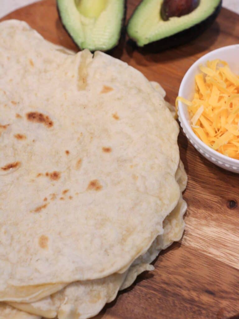 Tortillas stacked on a cutting board with a bowl of cheese and avocado in the background. 