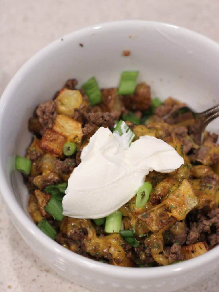 potatoes, meat and cheese in a white bowl ready to eat topped with sour Cream and green onions. 