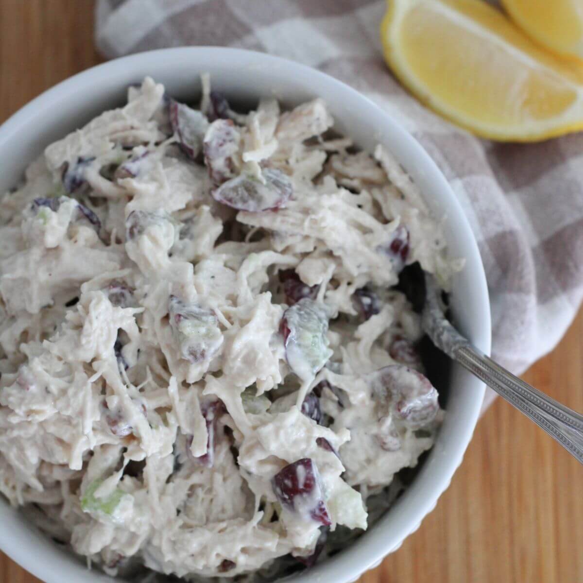 High- Protein Chicken salad in a white bowl with a silver spoon on a cutting board with a plaid napkin next to it and a cut lemon