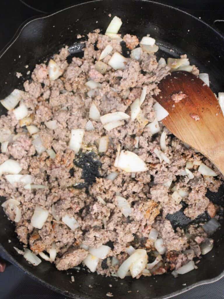 Browning the ground beef with garlic, onion, and seasonings in a cast-iron pan with a wooden spoon 