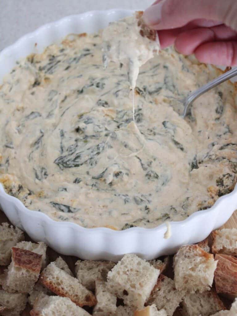 close up of the spinach dip dripping off a piece of sourdough bread above the glass serving bowl and sourdough bread around it. 