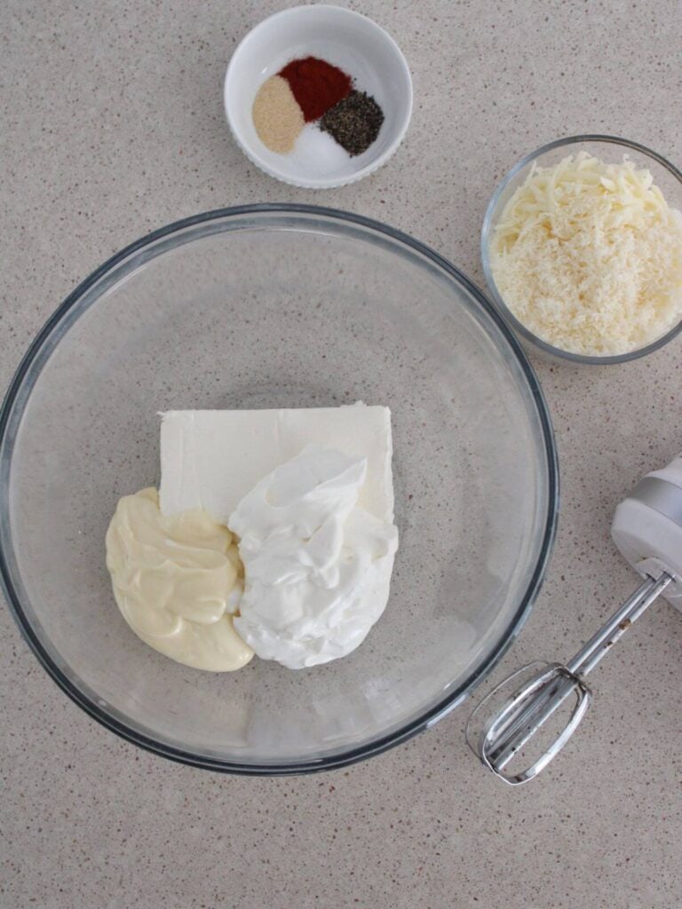 the cream cheese, sour cream and mayo being mixed together in a glass bowl with cheese and spices around and a hand mixer