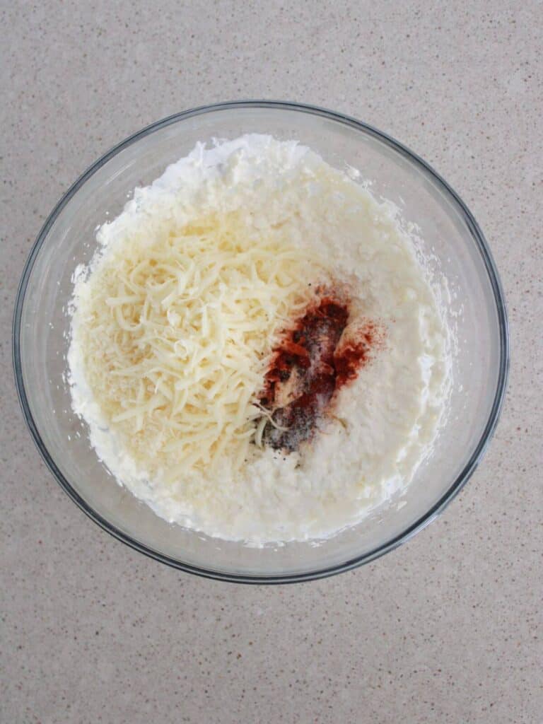 cheese and seasonings being mixed into the sour cream, mayo and cream cheese base in a glass bowl